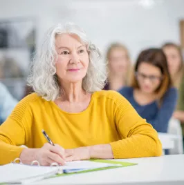 A diverse group of students are indoors in a university. They are taking night classes. A Caucasian senior woman is in front, and she is smiling while watching the lecture.